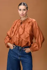 Camisa de color terracota con cuello mao, cierre frontal de botones y volados verticales a lo largo de la pechera. Presenta mangas largas abullonadas con puños ajustados.