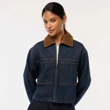 Chaqueta vaquera de color azul oscuro, corte corto y holgado, con cierre frontal de cremallera y dos bolsillos grandes con solapa en el frente. Posee cuello de pana marrón.