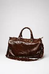 Bolso de viaje color marrón, confeccionado en cuero vacuno, con doble asa de mano y correa ajustable y desmontable. Cuenta con herrajes metálicos y costuras reforzadas.