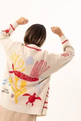 Saco tipo cardigan oversize de punto color beige, con diseño temático marino que incluye bordados de corales, peces, estrellas de mar y tortugas. Presenta detalles de galón picot en color rojo en los bordes y puños, cierre frontal con botones al tono y corte holgado.