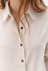 Camisa beige de manga larga con botones marrones y cuello clásico. Presenta un detalle de costura dorada a lo largo de la botonera.
