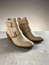Bota corta de cuero natural color beige, con taco de madera, puntera afinada y cierre trasero. Cuenta con tiras con hebillas metálicas y detalles pintados a mano en la parte trasera.