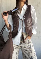 Chaleco de gamuza color chocolate con bordados decorativos en color crudo y cierre frontal con cordones y borlas.
