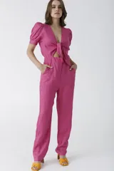 Mono fucsia de lino con escote en V profundo, nudo frontal que deja ver un corte en la cintura y mangas cortas abullonadas. El pantalón es de corte recto y holgado.