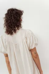 Vestido midi color crudo, con cierre frontal de botones, escote en V y mangas cortas abullonadas. Presenta un diseño holgado con detalle de volados en el bajo.