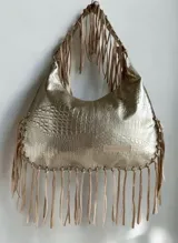 Cartera de hombro color dorado con textura símil cocodrilo y flecos colgantes en los bordes y las asas, de la marca Carolina de Cupto.