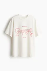 Remera de algodón color crema, con cuello redondo y mangas cortas. Presenta un estampado central en rojo con tipografía cursiva que dice "Papaya", y texto adicional que incluye "Beach Cafe", "Half Moon Bay", "Est 1985" y "Antigua", junto a un pequeño gráfico de una papaya.