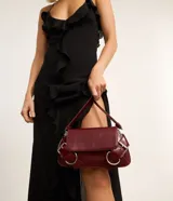 Cartera baguette Satinato pequeña de color negro, confeccionada en poliuretano. Presenta una solapa frontal con cierre de botón a presión, correa ajustable para el hombro y detalles decorativos de hebillas con argollas metálicas plateadas en los laterales.