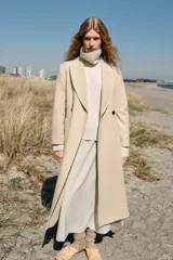 Abrigo largo de lana en color beige, con cuello de solapa, cierre cruzado con botones y cinturón ajustable del mismo tejido. Presenta mangas largas con detalle en los puños y bolsillos de vivo en el delantero.