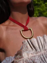 Collar con dije dorado en forma de estribo y tira de cuero rojo, de diseño artesanal con inspiración ecuestre.