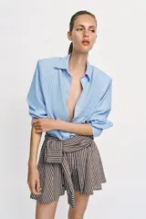 Bermuda de tiro medio con estampado de rayas verticales en tonos marrones y celestes. Presenta un diseño único con lazada frontal que simula mangas anudadas, elástico en la parte posterior de la cintura, cierre frontal con cremallera y botones, y bajo con vuelo.