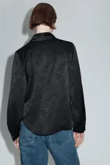 Camisa de satén negro con diseño jacquard de flores tono sobre tono. Presenta escote en pico con solapas, cierre frontal con botones y un broche metálico decorativo en el centro. Tiene mangas largas con puños abotonados.