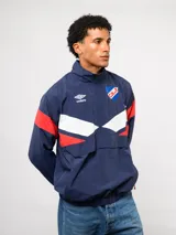 Buzo deportivo de nylon con medio cierre frontal y cuello alto. Presenta un diseño de bloques de color en azul, blanco y bordó, con el escudo del Club Nacional de Football y el logo de Umbro en el pecho.