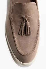 Mocasines de gamuza color marrón topo, con borlas decorativas en el empeine, costura frontal tradicional y suela plana de goma en color blanco off-white.