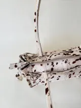 Cartera de cuero de tamaño medio con estructura rígida. Presenta una tira decorativa con tachas y hebillas metálicas en los extremos, además de lazos laterales. Cuenta con doble asa para llevar al hombro y cierre superior.