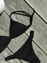 Conjunto de bikini negro con acabado brillante y textura sutil. El top es de estilo triángulo fijo con breteles regulables y cierre de gancho en la espalda. La bombacha es de corte vedetina con laterales fruncidos.