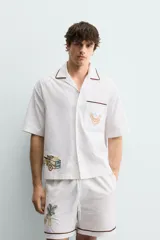 Camisa de corte relajado confeccionada en algodón, con cuello bowling y manga corta. Presenta un bolsillo en el pecho con vivo a contraste y bordados decorativos en el frente. Cierre frontal mediante botones.