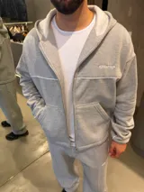 Campera con capucha gris de corte boxy, con cierre frontal completo, bolsillos tipo canguro y ribetes blancos en el pecho y las mangas. Cuenta con un pequeño estampado en el pecho.