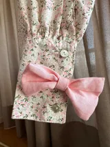Blusa de cuello alto con cierre de botones en la espalda, decorada con una hilera vertical de lazos rosados en el frente. Presenta mangas largas abullonadas con puños ajustados y lazos decorativos en los codos. Confeccionada en una tela con estampado floral de pequeñas flores rosas y verdes sobre fondo claro.