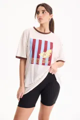 Remera de algodón blanca de manga corta, con corte suelto (loose fit) y rib en contraste marrón en el cuello y los puños. Presenta un estampado frontal rectangular con rayas verticales en tonos burdeos y celestes, superpuesto con el dibujo de una rodaja de limón amarilla y texto impreso.