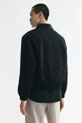 Campera negra de sarga de algodón, con cuello inglés, cierre frontal metálico y bolsillos delanteros ribeteados. Presenta elástico revestido en puños y bajo, y diseño forrado en su interior.
