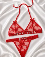 Conjunto de lencería rojo confeccionado en tul bordado, compuesto por un soutien triangular con tirantes finos para anudar al cuello, moños decorativos y aplicaciones de letras brillantes con la frase "TODA TUYA", y una bombacha colaless con la palabra "DANI".