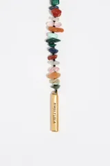 Collar estilo bolo o lariat, compuesto por piedras semipreciosas de diferentes colores y formas irregulares, ensartadas en dos hebras que se unen mediante un anillo central dorado. Las hebras cuelgan libremente y terminan en tubos metálicos dorados.