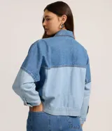 Campera de jean con diseño bicolor, confeccionada en algodón. Presenta cuello polo, cierre frontal con botones, mangas largas y bolsillos aplicados en el frente con recortes estructurados.