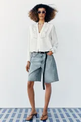 Blusa blanca de manga larga con cuello en V, cierre frontal con botones y detalles de bordados florales y encaje en el frente.