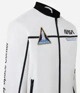 Campera tipo bomber blanca con detalles en negro, inspirada en uniformes espaciales de la NASA. Presenta parches con el logo de la NASA, la bandera de EE. UU. y texto en la manga que dice "Kennedy Space Center".