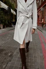 Abrigo de paño negro con capucha, cierre frontal cruzado con botones y cinturón de lazada en el mismo tejido. Presenta manga larga con trabillas en los puños y bolsillos laterales.