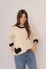 Buzo de punto tejido a mano 100% lana merino, color beige con detalles en negro en el cuello, los puños y un diseño de corazón en el borde inferior. Su diseño es amplio de manga larga.