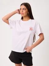 Remera blanca de corte holgado, con cuello redondo y mangas cortas. Presenta un pequeño estampado en el pecho con diseño floral y tipografía.