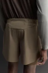 Short de running 2 en 1 color beige, confeccionado en tejido técnico ligero y elástico con malla interior integrada. Presenta cintura elástica ajustable con cordón, bolsillo trasero con cierre y aberturas laterales en el bajo.