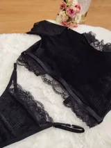 Conjunto de lencería compuesto por un top de cuello alto y una tanga, confeccionados en terciopelo negro con detalles de encaje y microtul. El top presenta mangas cortas con terminaciones en encaje y el borde inferior decorado con una cinta y encaje. La tanga es regulable y combina la textura del terciopelo con bordes de encaje.
