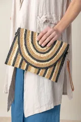 Sobre o clutch de rafia con diseño de franjas semicirculares en tonos naturales (beige claro, dorado con lurex, marrón y negro). Presenta un borde trenzado y correa de mano de cuero marrón.