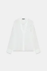 Blusa de tejido semitransparente con efecto arrugado, escote en pico y manga larga. Presenta un detalle decorativo frontal con estructura en forma de flor.