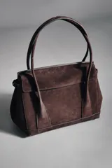 Bolso tipo citybag confeccionado en serraje color marrón oscuro. Presenta una solapa frontal con cierre magnético, asas de hombro integradas y detalles de hebillas metálicas en los laterales para ajuste. Incluye bolsillo interior con cremallera.