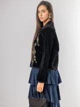 Chaqueta de terciopelo negro con bordados florales y ornamentales en tonos dorados, rosados y azules. Presenta un corte recto, cuello redondo y cierre frontal abierto con detalles de costuras decorativas en los bordes.