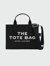 Bolso tipo tote de lona en color negro con el nombre del modelo y la marca estampados en blanco en el frente. Cuenta con doble asa superior, correa de hombro ajustable y extraíble, y cierre superior con cremallera.