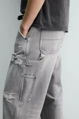 Pantalón de jean estilo carpintero con corte relaxed fit, tiro medio y pierna recta. Presenta un lavado azul medio con efecto desgastado y roturas en las rodillas. Incluye bolsillos laterales tipo plastrón, tira multifunción en la pierna, cierre frontal con cremallera y botón.