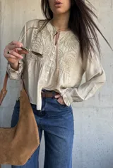 Camisa beige de manga larga con detalles bordados en el frente, cuello redondo y cierre frontal con botones. Las mangas presentan puños con volados.