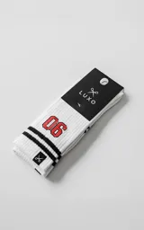 Medias blancas de caña alta con diseño acanalado, detalle de dos franjas negras horizontales y el número 06 bordado en color rojo. Incluye etiqueta pequeña con logo de tijeras en el borde superior.