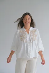 Blusa blanca de corte holgado con cuello mao y cierre frontal de botones. Presenta mangas largas abullonadas con puños elásticos y un delicado bordado en color beige en la parte superior delantera.