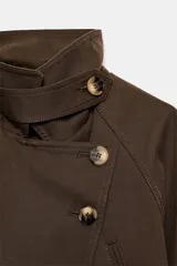 Cazadora trench corta color marrón, confeccionada en mezcla de algodón, con cuello solapa, cierre cruzado con botones, bolsillos de vivo en el delantero y cinturón ajustable del mismo tejido.