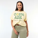 Remera de corte oversize en color amarillo claro, con cuello redondo y mangas cortas. Presenta un estampado gráfico frontal con la frase 'COSTA AZUL' en letras verdes y un diseño de sol, acompañado por el texto 'Palma de Majorque' en tipografía cursiva.