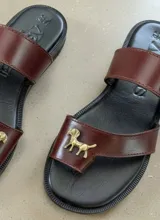 Sandalias de cuero color marrón con base negra, diseño de doble tira y aplique metálico de bronce con forma de perro.