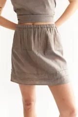 Falda corta de lino con cintura elástica y pliegues verticales. El tejido es de color base gris/marrón con finas rayas verticales en tono ocre/amarillento.