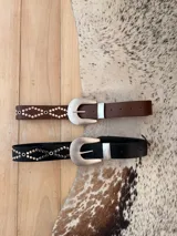 Cinturon de cuerina negra con tachas plateadas dispuestas en patron geometrico y hebilla metalica grande de estilo western.