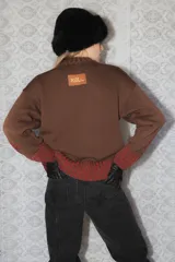 Buzo de tejido de punto grueso color marrón con diseño geométrico y floral estampado en color rojo. Presenta cuello redondo, mangas largas y puños y ruedo acanalados.